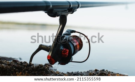 Spinning rod on shore near water outdoors, close-up of steel red reel. Fishing hobby, sport and leisure concept. Selective soft focus. Royalty-Free Stock Photo #2115355598