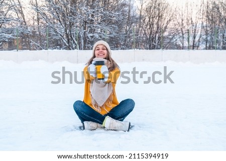 Portrait of happy young woman in yellow sweater and ice skates sitting on the snow and holding out mug with coffee to camera.