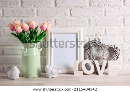 Empty photo frame with bouquet of pink tulips, word love and birds on the white brick wall. Romantic spring background with space for text.