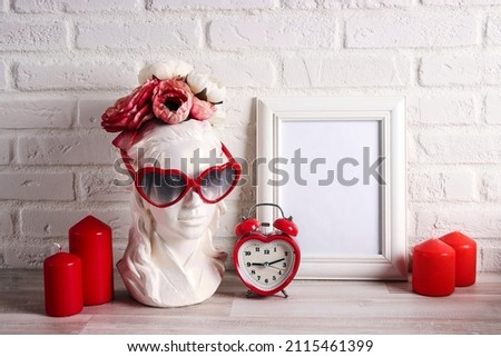 Empty photo frame with female plaster head with heart shaped glasses, flowers, alarm clock and candles against the white brick wall. Copy space. Romantic and love, Valentine's Day concept.
