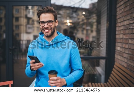 Half length of funny hipster guy in classic spectacles holding modern cellphone gadget and takeaway caffeine beverage and smiling at camera during weekend leisure in city, happy male blogger posing Royalty-Free Stock Photo #2115464576