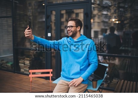 Happy Caucasian man using front mobile camera on modern cellular technology for making selfie content during weekend leisure, cheerful male millennial shooting video vlog via smartphone application