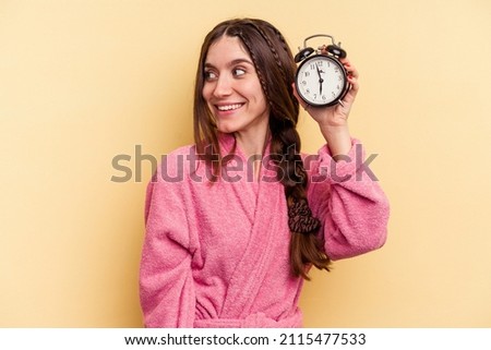Young caucasian woman wearing a bathrobe holding a alarm clock isolated on yellow background looks aside smiling, cheerful and pleasant.