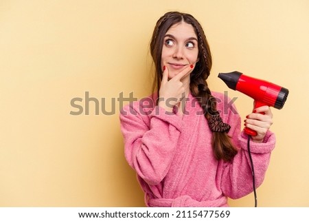 Young woman holding a hairdryer isolated on yellow background looking sideways with doubtful and skeptical expression.