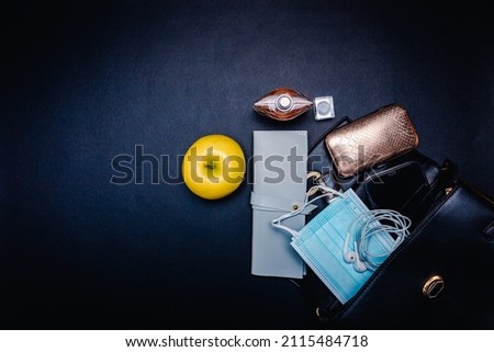 Daily accessories coming out of woman's bag, masks, perfume, smartphone, apple and other on black background