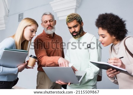 Middle aged teacher with cellphone standing near multiethnic students with laptops in university corridor Royalty-Free Stock Photo #2115509867