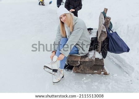 Lovely young woman sitting on the bench and puting on figure skates on the ice rink. Girl is going to skate on ice in a winter frosty day
