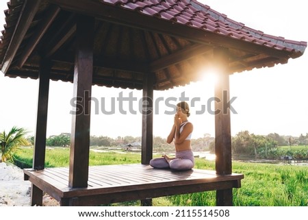 Side view of calm female in sportive tracksuit sitting in lotus pose and keep praying during morning meditation near rice fields, young Caucasian woman relaxing during yoga concentration in Thailand Royalty-Free Stock Photo #2115514508
