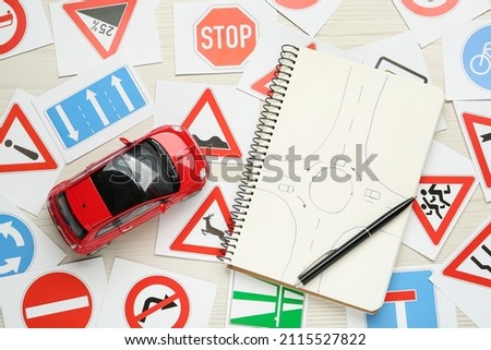 Many different road sign cards, notebook with sketch of roundabout and toy car on white wooden background, flat lay. Driving school