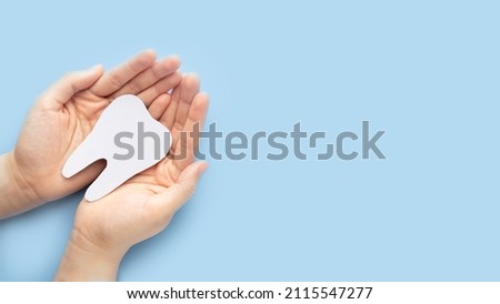 Woman hand holding healthy white teeth symbol on light blue background. Dental care, tooth protection and oral hygiene concept. Top view. Copy space.