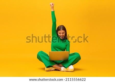 Winner. Excited happy student teen girl sitting on floor with laptop, raising one hand in the air if saying yes, isolated on yellow background
