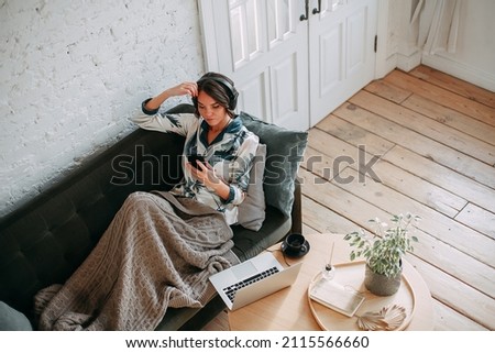 Young brunette woman working remotely from home. She sits on a dark green sofa in black headphones and looks at the smartphone screen.