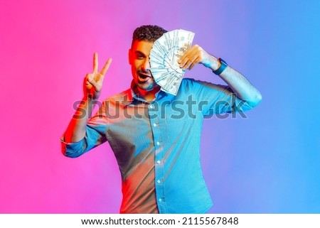 Portrait of rich funny man in shirt covering half of face with dollars, looking at camera showing vi sign, being happy to win lottery. Indoor studio shot isolated on colorful neon light background.