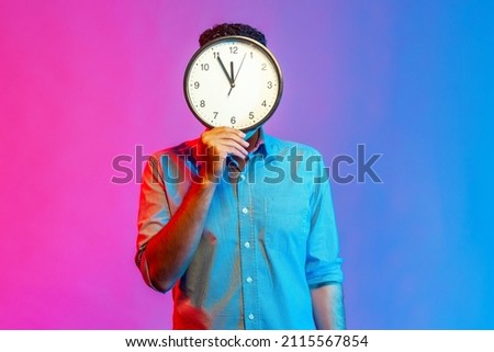 Portrait of man in shirt hiding face behind big wall clock display, wasting his time, procrastination, bad organization of working time. Indoor studio shot isolated on colorful neon light background. Royalty-Free Stock Photo #2115567854