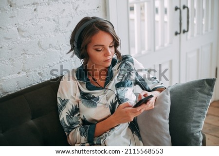 Young woman working remotely from home. She sits on a dark green sofa in black headphones and looks at the smartphone screen.