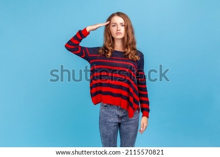 Yes sir Portrait of serious woman wearing striped casual style sweater, standing saluting with hand near head, looking obedient and attentive at camera. Indoor studio shot isolated on blue background Royalty-Free Stock Photo #2115570821