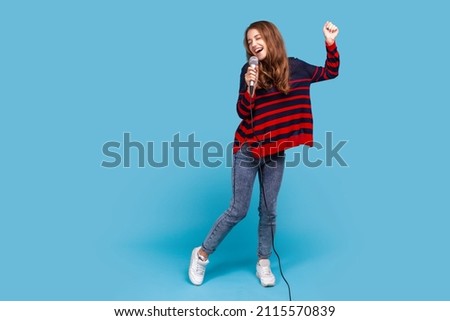 Full length portrait of woman wearing striped casual style sweater, singing in microphone and dancing, having fun in karaoke, performing alone. Indoor studio shot isolated on blue background.