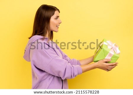 Side view portrait of smiling young adult woman holding out green wrapped gift box, giving present, congratulating, wearing purple hoodie. Indoor studio shot isolated on yellow background.