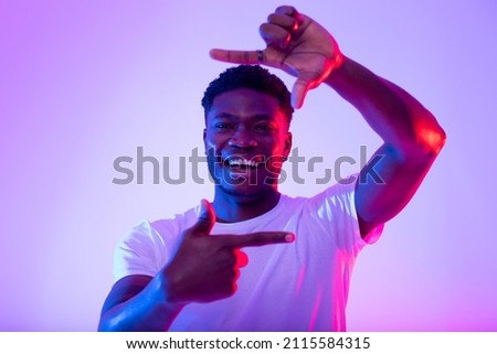Cheerful young black man making picture frame with fingers, looking at camera and smiling for photo in neon light. Handsome African American guy showing photograph gesture. Capturing moments