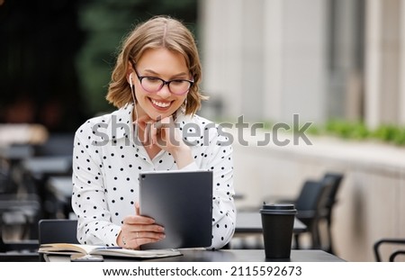 Happy young enthusiast female teacher tutor freelancer in earphones sitting in front of tablet on terrace outside emotionally gesticulating with bright smile explains to students during online lesson Royalty-Free Stock Photo #2115592673