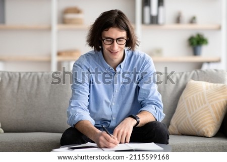Positive male student with notebook sitting on sofa at home and studying remotely, making homework, taking notes, getting ready for exam, free space