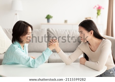 Mature Asian mother and adult daughter arm wrestling, looking each other in eyes, fighting at home. Parent child disagreement, confrontation between mother-in-law and daughter-in-law concept