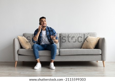 Making Call. Portrait Of Cheerful Handsome Arab Man Talking On Smart Phone Sitting On Sofa At Home In Living Room, Having Pleasant Conversation With Friends Or Family, Looking Away Gesturing Royalty-Free Stock Photo #2115599303