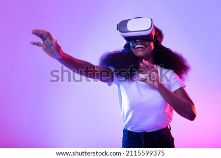 Happy young black lady in VR glasses exploring cyberspace, touching imaginary screen, playing virtual reality game in neon light. Positive African American woman experiencing contemporary technologies Royalty-Free Stock Photo #2115599375