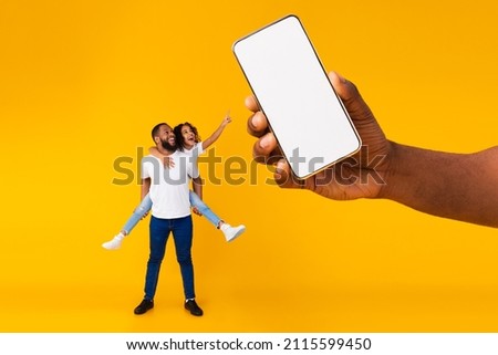 Great Offer. Cheerful black dad carry daughter kid on back and shoulders, excited girl pointing at giant mobile phone showing blank space for mock up, hand holding white empty screen, full body length Royalty-Free Stock Photo #2115599450