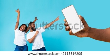 Great Offer. Cheerful African American couple dancing and pointing at giant cell phone showing blank space for mock up, celebrating win, hand holding white empty screen, blue studio background wall Royalty-Free Stock Photo #2115619205