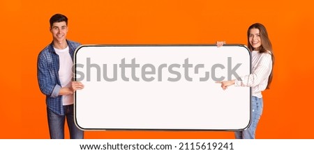 Great Offer. Portrait of excited couple holding big horizontal smartphone with white blank screen in hands and pointing at device. Gadget with empty free copy space for mock up, banner, panorama Royalty-Free Stock Photo #2115619241