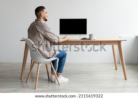 Rear Back View Of Happy Smiling Arab Man Looking At Pc Computer With Blank Black Screen For Mock Up Template Sitting At Desk At Home Office, Free Copy Space. People, Technology, Remote Work Concept Royalty-Free Stock Photo #2115619322