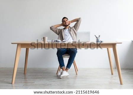 Taking Break From Work. Portrait of smiling young Arab man relaxing on chair sitting at table and using laptop, happy millennial male leaning back at workplace and dreaming, holding hands behind head Royalty-Free Stock Photo #2115619646