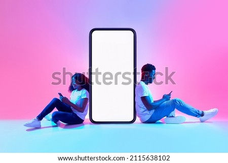 Cool mobile app. African American couple sitting near big smartphone with mockup, using modern gadgets, advertising new application or website, promoting your product or service in neon light Royalty-Free Stock Photo #2115638102