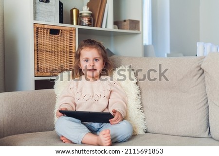 School online. Adorable little girl of 3 years old is sitting on the couch in a cozy house and talking on a video call. Portrait of an emotional child holding a tablet in his hands. Home.