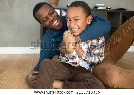 Funny picture of caring and loving dark-skinned dad hugging his misbehaving, naughty boy, both having fun, laughing, spending leisure time enjoying bonding and warm relationships, sitting on floor