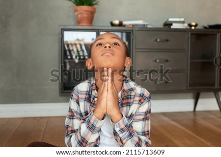 Indoor picture of handsome little man sitting on floor in praying position, keeping palms pressed together, looking up, making wish, asking God for help, luck. Body language, signs, gestures