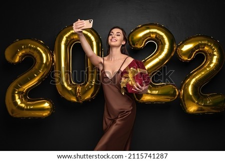 Smiling young brunette woman in elegant dress doing selfie on mobile phone, black background golden numbers air balloons. Holding red gift box. Happy New Year 2022 celebration holiday party concept.