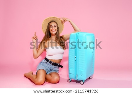A young woman in a hat, denim shorts and white top while sitting near a blue suitcase pointing to empty space. Pink studio background