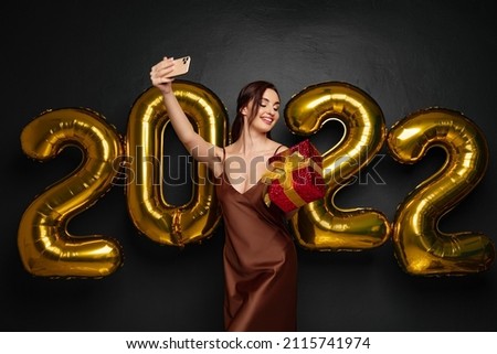 Smiling young brunette woman in elegant dress doing selfie on mobile phone on black background golden numbers air balloons. Holding red gift box. Happy New Year 2022 celebration holiday party concept.