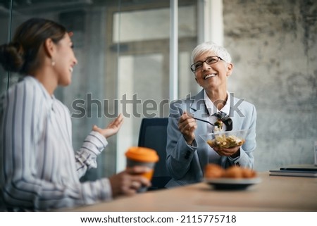Happy mature businesswoman enjoying in conversation with female coworker during her lunch break in the office. Royalty-Free Stock Photo #2115775718