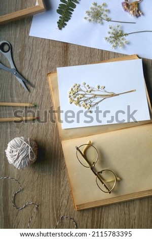 Old book, papers, various pressed flowers, eyeglasses, scissors, pencils and rope on wooden desk. Crafting and making herbarium at home. Flat lay.