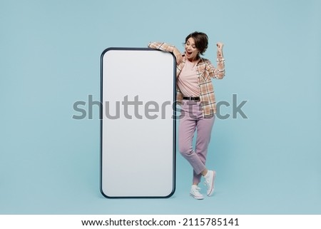 Full body young smiling happy woman 20s in casual brown shirt stand near big mobile cell phone with blank screen workspace area do winner gesture isolated on pastel plain light blue color background. Royalty-Free Stock Photo #2115785141