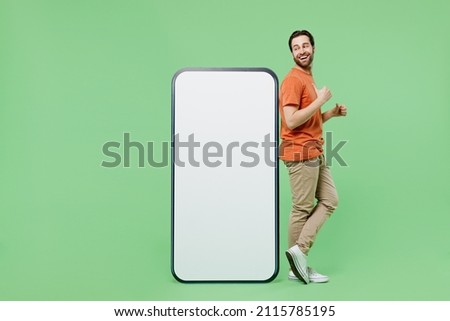 Full size fun young man 20s wear orange t-shirt stand point finger on mobile cell phone with blank screen workspace area isolated on plain pastel light green color background People lifestyle concept