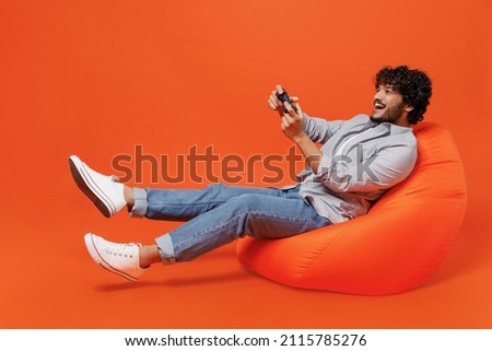 Full size body length vivid young bearded Indian man 20s years old wears blue shirt sit in bag chair hold in hand play pc game with joystick console isolated on plain orange background studio portrait