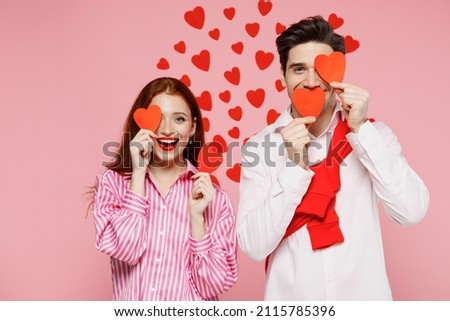Young cheerful couple two friends woman man in casual shirt hold paper red hearts cover eyes isolated on plain pastel pink background studio portrait. Valentine's Day birthday holiday party concept