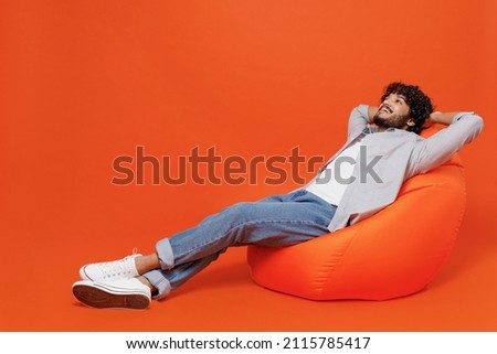 Full size body length joyful excited fancy young bearded Indian man 20s wears blue shirt sit in bag chair put his hands behind head resting chilling isolated on plain orange background studio portrait