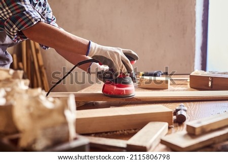 Female carpenter grinding wood with sandpaper in carpentry or diy workshop. Electric sander working in carpentry. Girl polishes wooden board with electric sander. Royalty-Free Stock Photo #2115806798