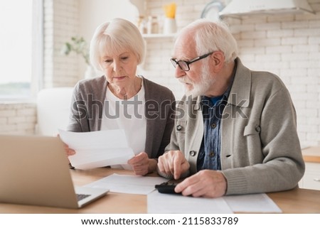 Serious caucasian old elderly senior couple grandparents family counting funds on calculator, doing paperwork, savings, paying domestic bills, mortgage loan, pension at home using laptop. Royalty-Free Stock Photo #2115833789