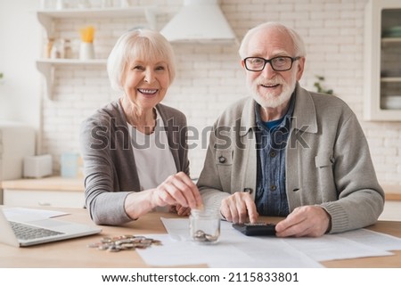 Savings concept. Nest egg of old elderly senior couple grandparents husband and wife saving money, economizing pension, mortgage loan at home using laptop and putting coin into moneybox